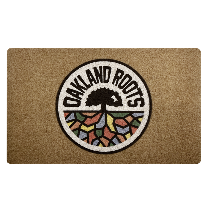 The Oakland Roots SC Door Mat by Oakland Roots SC showcases a circular logo depicting a stylized tree with roots resembling vibrant, mosaic-like city blocks. The text "OAKLAND ROOTS" surrounds the tree in bold, black letters on a white background, reflecting the team's commitment to social good.