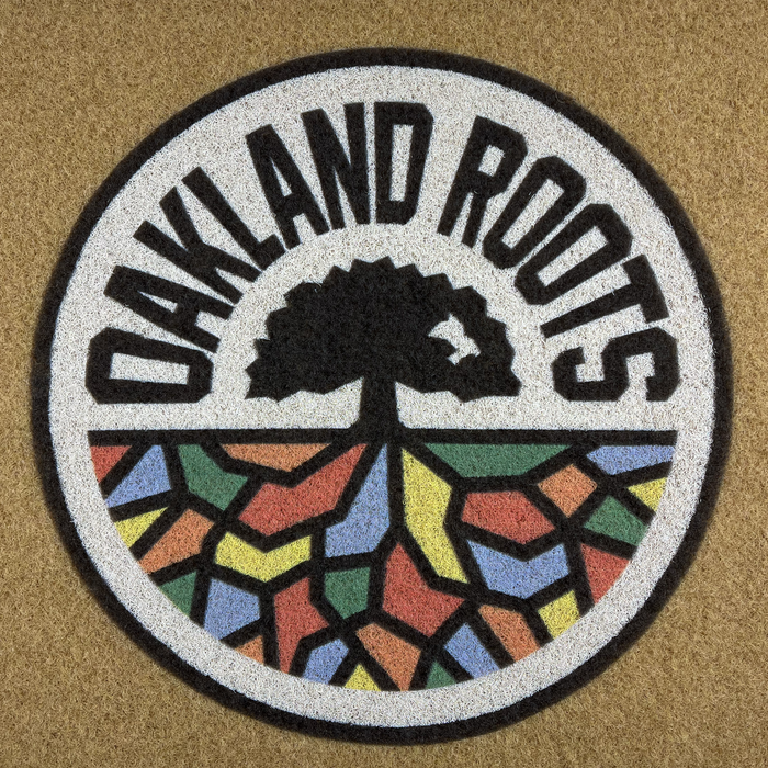 The image shows the logo of the Oakland Roots SC, as featured on the Oakland Roots SC Door Mat. The logo depicts a black tree with visible roots inside a circle. Arched over the top of the circle is the text "OAKLAND ROOTS." The lower half of the circle is filled with vibrant geometric shapes, reminiscent of stained glass, symbolizing social good.