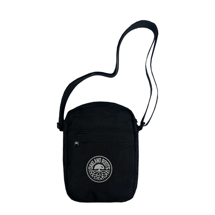 The Oakland Roots SC Flight Bag is a black crossbody with a single shoulder strap and a front zippered pocket. It features a white circular logo that reads "OAKLAND ROOTS" encircling a tree with roots, symbolizing the sports club and organization dedicated to social good. The bag boasts a minimalist, casual design.