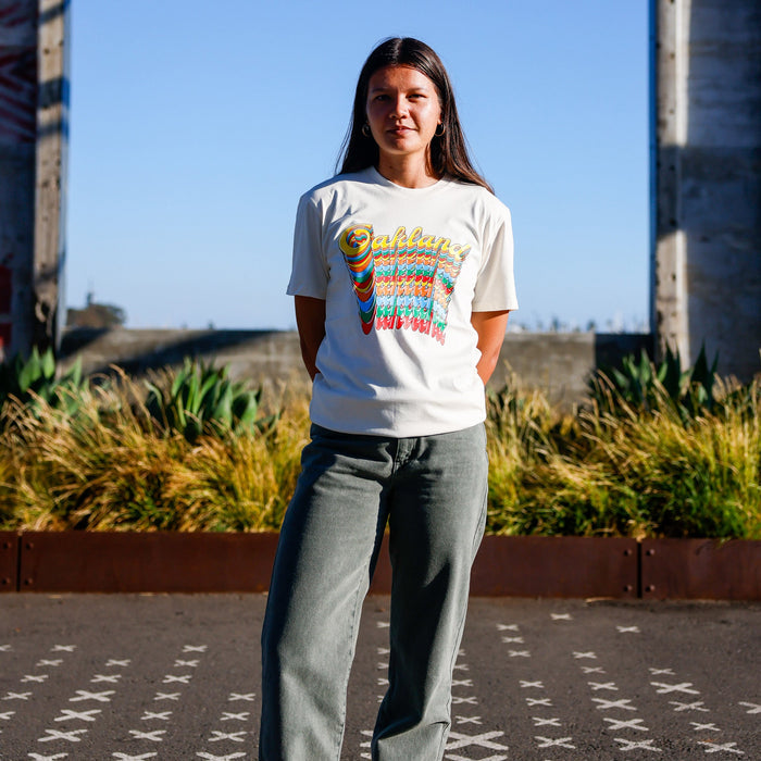 A woman with long dark hair stands outdoors on a sunny day, hands behind her back. She wears the Oakland Roots SC Funk Tee by Other, featuring colorful “Oakland” text, paired with gray-green pants. Plants and tall concrete structures are in the background beneath a clear blue sky.