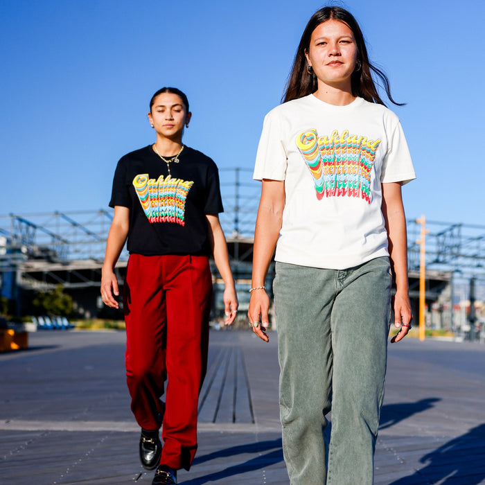 Two women walk outdoors on a sunny day. The woman in front sports a white Oakland Roots SC Funk Tee by Other and green pants, while the woman behind wears a black Oakland t-shirt and red pants. In the background, a wooden walkway and metal structures add to the urban setting.