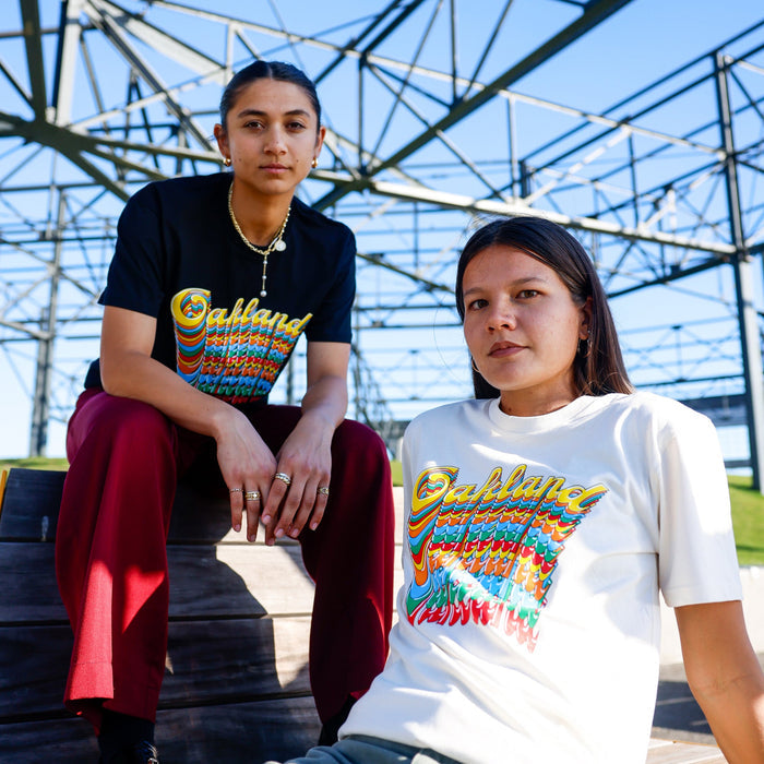 Two people sit outdoors on wooden steps beneath a metal structure. One wears the black Oakland Roots SC Funk Tee by Other with red pants, while the other wears a white “Oakland” shirt and light pants. Both face the camera with serious expressions under a blue sky.