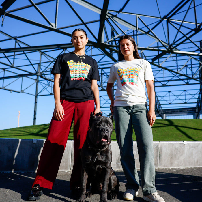 Two people stand side by side under a metal structure, both wearing graphic t-shirts—including the Oakland Roots SC Funk Tee by Other—and casual pants. A large black dog sits in front of them. The sky is clear and blue, and green grass is visible in the background.