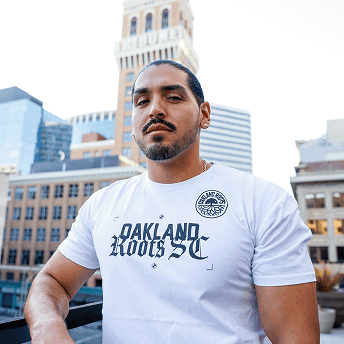 A man with dark hair pulled back and a goatee stands on a city rooftop, wearing the Oakland Roots SC Futbol Town Tee by Other. Behind him, tall buildings and a historic clock tower rise against a clear blue sky.