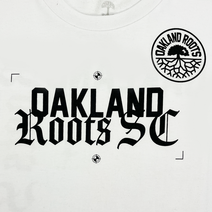 The Oakland Roots SC Futbol Town Tee by Other features a bold, black gothic “Oakland Roots SC” print on a white shirt. The design includes a circular Oakland Roots Sports Club logo above the text, showcasing a tree with roots and the team name. Small target marks highlight the text’s corners and center for added detail.