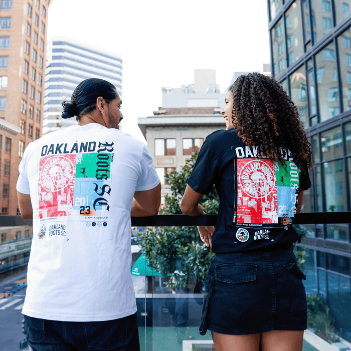 Two people stand on a glass balcony overlooking the city, both sporting the Oakland Roots SC Futbol Town Tee by Other. One wears the tee in white and the other in black, each featuring a vibrant graphic design on the back. The tall buildings and greenery in the background highlight their bold shirts against the lively urban scene.