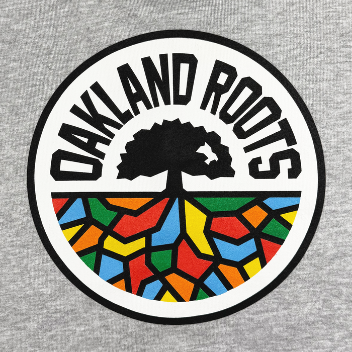 A circular logo on a gray background features the bold black text "Oakland Roots SC" from the Oakland Roots SC brand, representing a dynamic sports club. Beneath it, a stylized black tree stands atop a vibrant geometric mosaic with sections in red, yellow, green, orange, and blue, symbolizing its dedication to social good.
