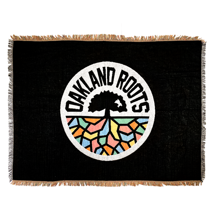 The Oakland Roots SC Throw is a rectangular black tapestry showcasing the Oakland Roots Sports Club logo in the center, which features a tree with sprawling roots above a vibrant, mosaic-like ground. The words "Oakland Roots" encircle the tree within a white-bordered circle. This socially conscious throw is accented with fringe along its edges.