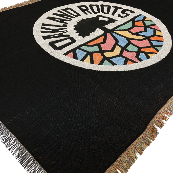 The Oakland Roots SC Throw by Oakland Roots SC is a woven black blanket adorned with a circular emblem. The center of the emblem showcases a tree, with "Oakland Roots Sports Club" arched above it. Below the tree are colorful geometric shapes in blue, red, orange, yellow, and green. The blanket is completed with multicolored fringe edges and radiates social good vibes.