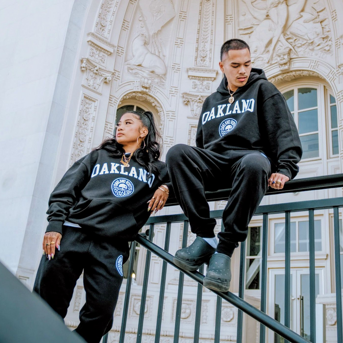 Two people in black Oakland Roots SC University Hoodies and sweatpants pose on metal railings before an ornate building. One stands, looking left; the other sits, looking down. Both wear jewelry and black shoes. Hoodies shown in men's sizing.