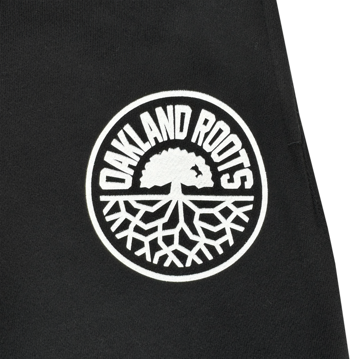 The Oakland Roots SC University Pant features black fabric with a white circular patch showing "OAKLAND ROOTS" above a stylized tree, symbolizing team pride, unity, and growth. Official gear by Oakland Roots SC.