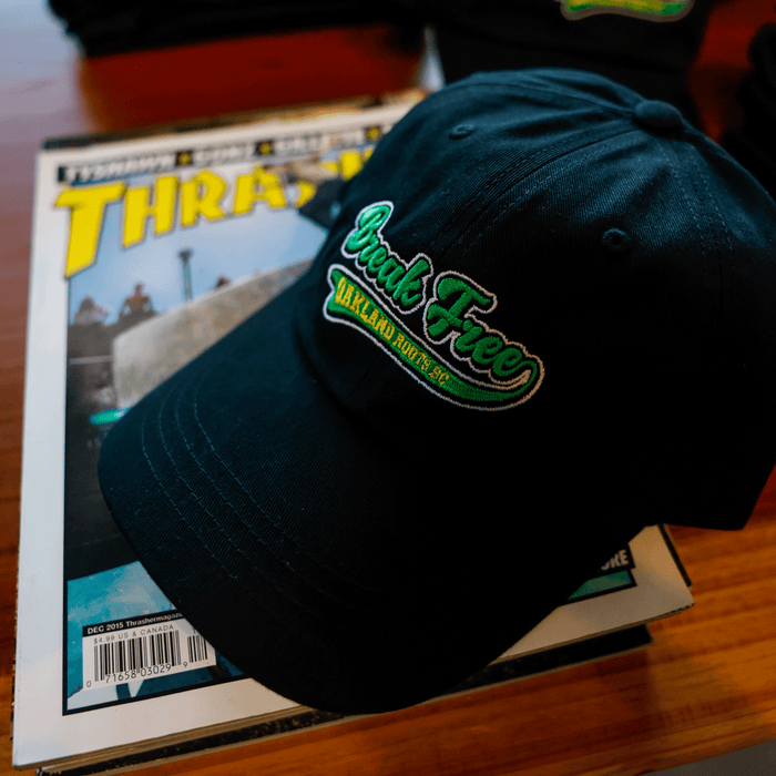 The Oakland Roots SC X Break Free Hat, a black dad hat with green and white embroidery, rests on a Thrasher skateboarding magazine atop a wooden surface. Its curved bill faces the viewer. Brand: Oakland Roots SC.