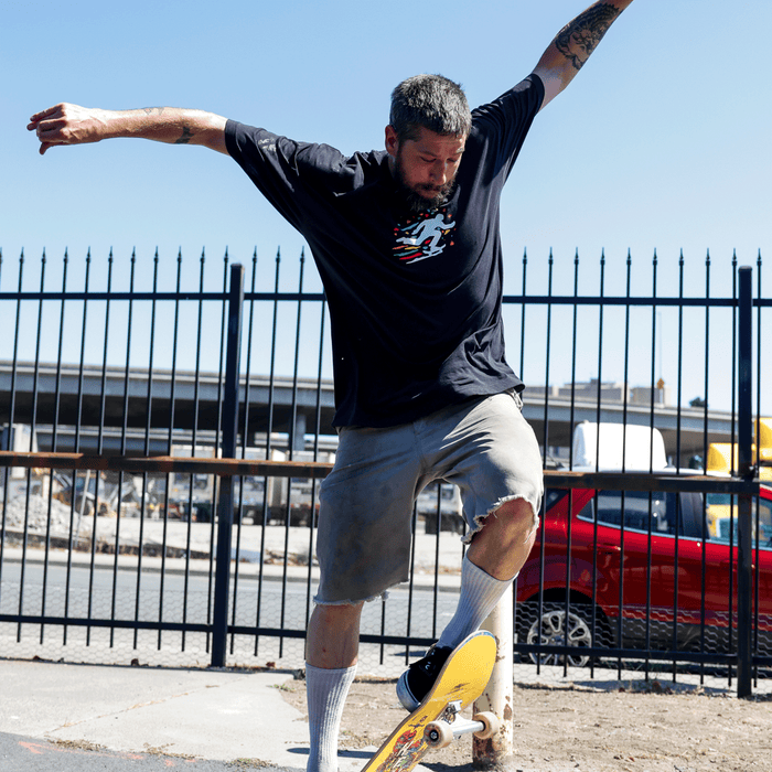 A tattooed, bearded man skateboards outdoors wearing the Oakland Roots SC X Break Free Tee by Oakland Roots SC, gray shorts, and white socks. He balances with arms outstretched as a red car and black metal fence reflect Oakland’s vibrant culture.