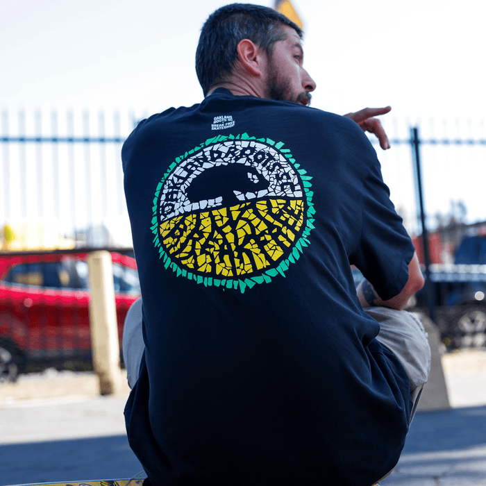 A man with short dark hair and a beard sits outdoors, facing away. He wears the Oakland Roots SC X Break Free Tee, featuring a colorful mosaic design inspired by Oakland culture. A fence and red car are blurred in the background.