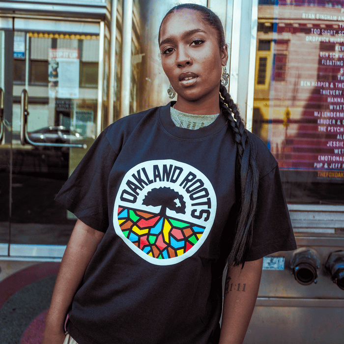A person with long braids and a visible tattoo confidently models the Oaklandish Oakland Roots SC X Oaklandish Bootleg Tee, standing before a venue window with event posters, showcasing the shirt’s colorful tree logo.