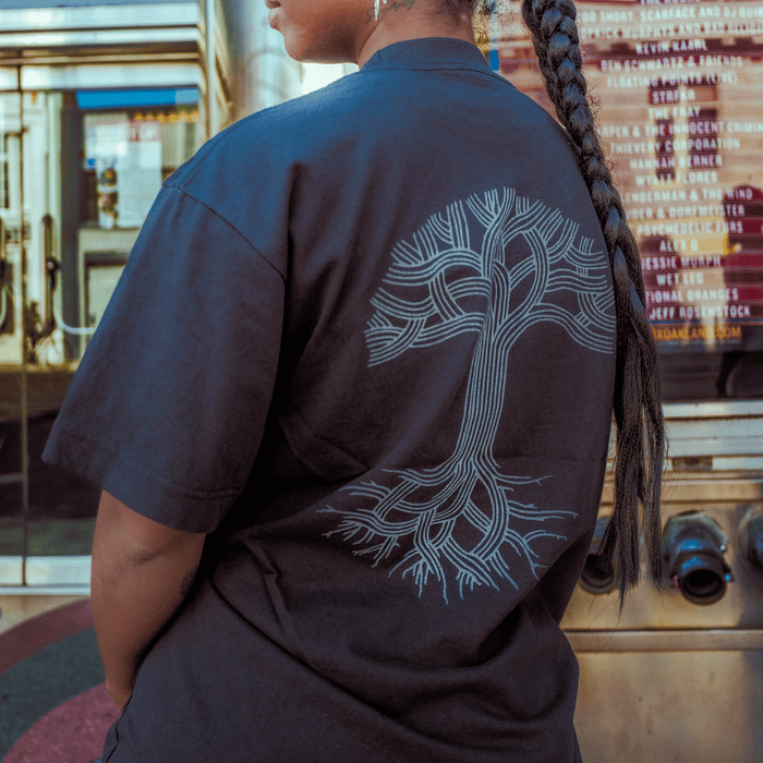 A person with a long braid wears an Oaklandish Oakland Roots SC X Oaklandish Bootleg Tee, which features a white tree design on the back. They stand by glass doors and a marquee with movie titles, suggesting an urban or theater location.