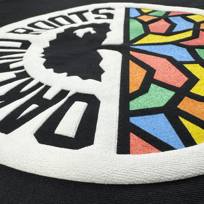 Close-up of textured black fabric on the Oakland Roots SC X Oaklandish Bootleg Tee by Oaklandish, featuring raised embroidery of a circular motif blending Africa’s silhouette and stained-glass patterns, with partial “ROOTS” text.