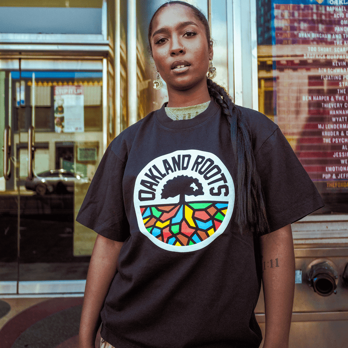 A person stands confidently outdoors in the Oaklandish Oakland Roots SC X Oaklandish Bootleg Tee, with long braids, hoop earrings, and a forearm tattoo. A glass building with posters and reflections is behind them.