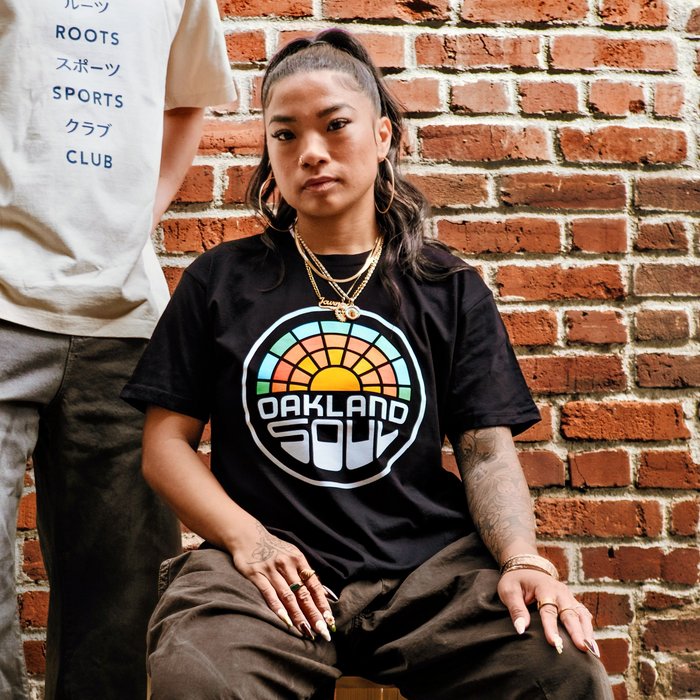 A woman with long dark hair in a ponytail sits by a brick wall, wearing the Oakland Soul Logo Minus Tee and brown pants—perfect for showing women's soccer pride. She accessorizes with hoop earrings, layered necklaces, and has tattoos.