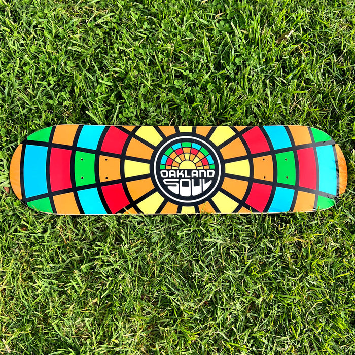 The Oakland Soul Logo Skate Deck, resting on the grass, showcases a vibrant geometric design. Constructed from durable 7-ply maple wood, it features a multicolored pattern in shades of red, orange, yellow, green, blue, and black arranged in a striking radial mosaic style. At the center is the Oakland Soul SC logo with “OAKLAND” in white above graffiti-style letters spelling “SOUL.”