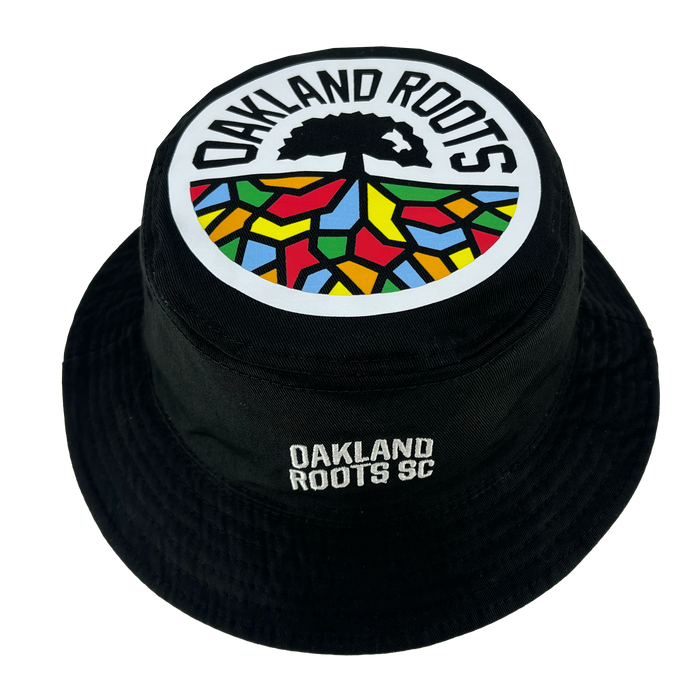 Black reversible bucket hat featuring the Oakland Roots SC logo on the front and top. The top logo includes a tree above a semi-circle with multicolored sections resembling stained glass. "OAKLAND ROOTS" arches over the top logo, and "OAKLAND ROOTS SC" is embroidered on the front. The product is called Oakland Soul & Roots SC Reversible Bucket by Oakland Roots SC.