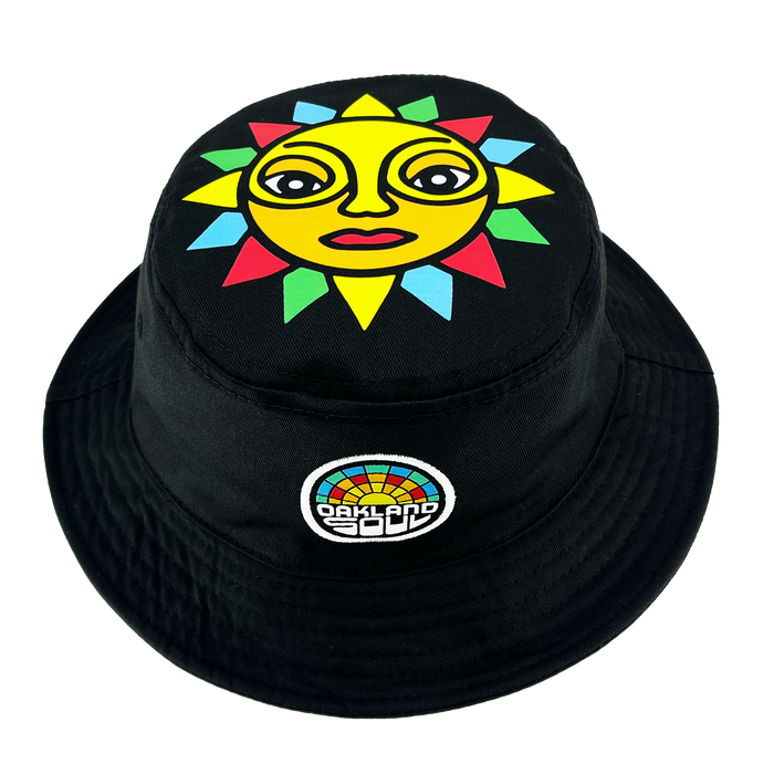 The Oakland Soul & Roots SC Reversible Bucket by Oakland Roots SC is a versatile black bucket hat that showcases a vibrant design on one side. The top of the hat features an illustration of a lively sun with a yellow face, expressive eyes, and multicolored rays. Accompanying the sun, there's a small patch adorned with a colorful semi-circle mosaic pattern and the text "Oakland Roots" below it.