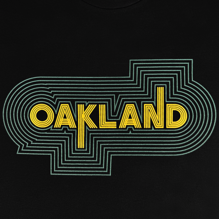 The image showcases an Oaklandish graphic featuring bold, yellow geometric "OAKLAND" text and multiple green concentric lines for a layered, retro sports vibe on the sleek black Oakland Sports Culture '68 Tee.