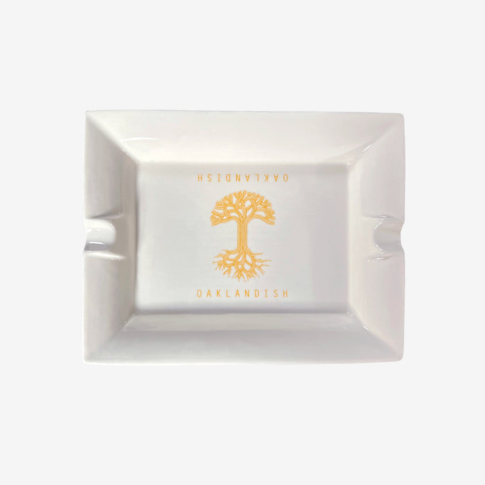 The Oaklandish Ceramic Ashtray by Oaklandish is a rectangular, white ceramic ashtray with a simple design showcasing an orange tree with visible roots in the center. The "OAKLANDISH" logo is imprinted above and below the tree in orange capital letters. Additionally, two grooves on opposite sides of the ashtray are designed to hold cigarettes.