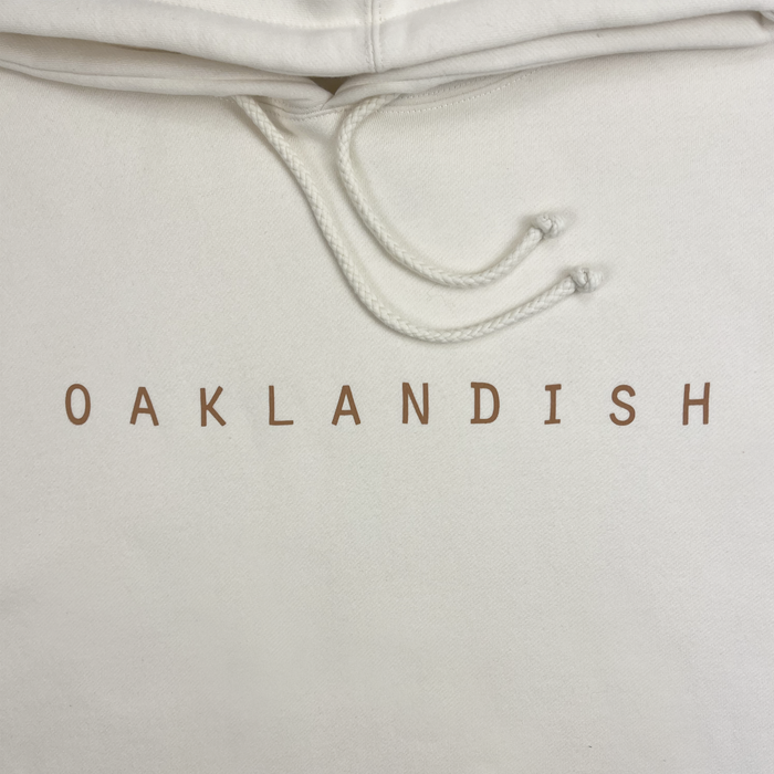 Close-up of the Oaklandish Classic Hoodie in white, featuring "OAKLANDISH" in bold brown, all-capital letters across the chest. This men's fit hoodie by Oaklandish has adjustable drawstrings tied in small knots at their ends, hanging from the hood. The fabric appears soft and well-finished.