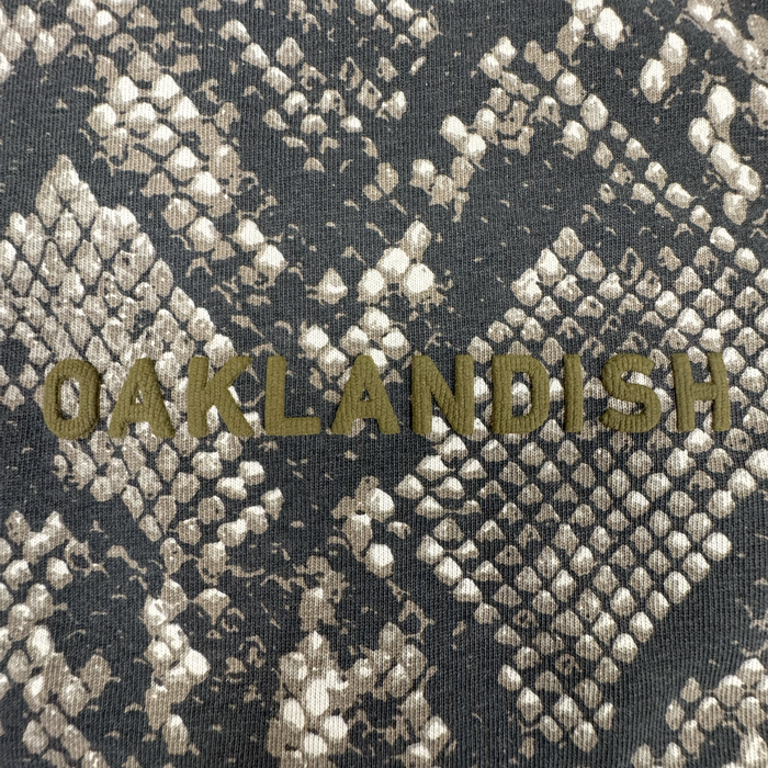 Crafted from 100% cotton for both comfort and style, the Oaklandish Classic Logo LS Tee by Oaklandish features a textured gray and tan snakeskin patterned background. Centered on the long sleeve t-shirt are bold, olive-green “OAKLANDISH” letters in an all-caps, sans-serif font. The design is complemented by vintage black sleeves for a unique look.