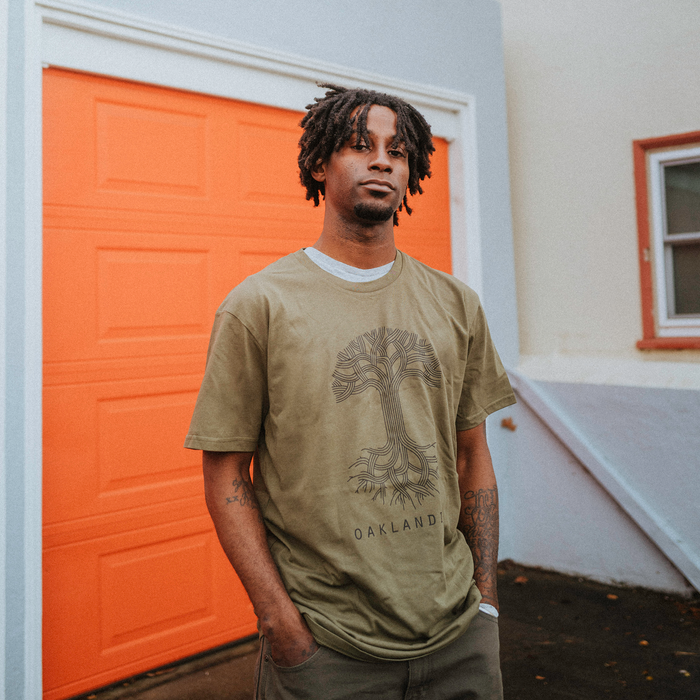 A man with short dreadlocks and a goatee stands before an orange garage door, wearing the green Oaklandish Classic Logo Tee featuring the iconic tree and "OAKLAND," paired with dark pants. A white building and small window are visible nearby.