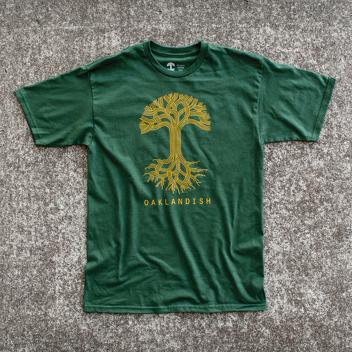 An Oaklandish Classic Logo Tee in dark green is laid flat on a gray surface. Made from 100% cotton, this shirt showcases a golden-yellow graphic of a stylized tree with intricate branching patterns above and below the ground. Below the tree graphic, matching golden-yellow text reads "OAKLANDISH.