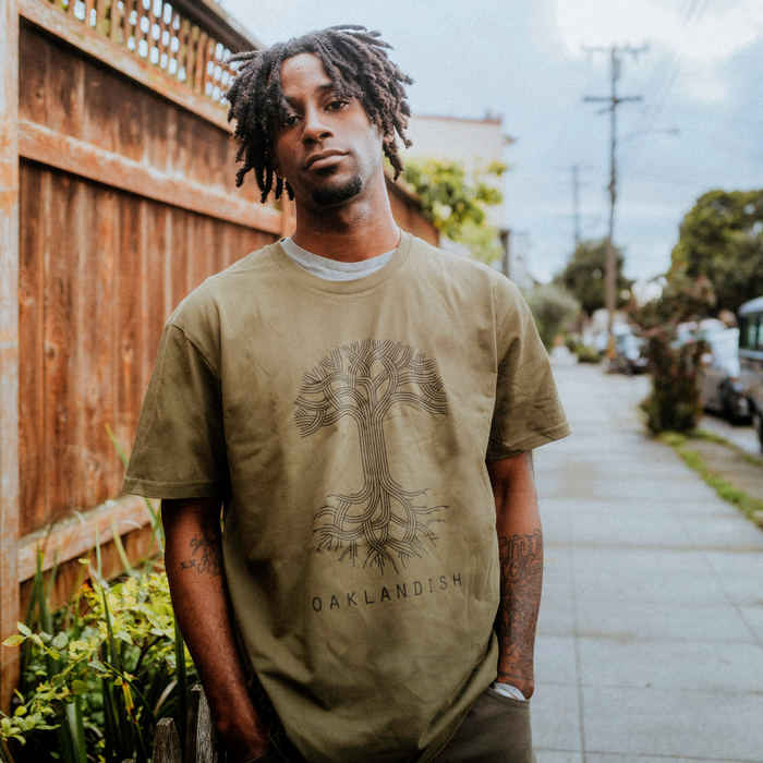 A young man with short dreadlocks stands on a sidewalk by a wooden fence, wearing the Oaklandish Classic Logo Tee. The tee features a classic tree graphic and bold "OAKLANDISH" text by the brand Oaklandish. Houses and greenery appear in the background.