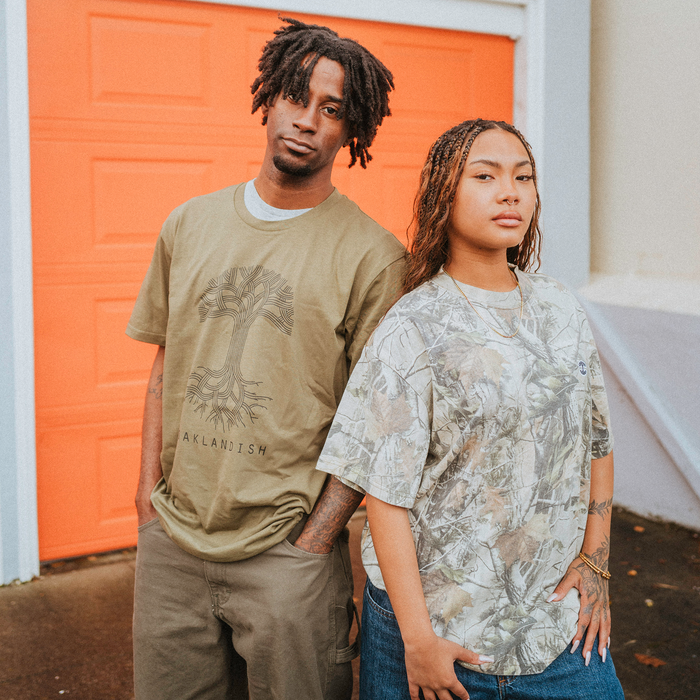 Two people stand by an orange garage door. On the left, one wears an Oaklandish Classic Logo Tee with khaki shorts; on the right, the other wears a loose light camo men's cotton shirt and jeans. Both look relaxed.