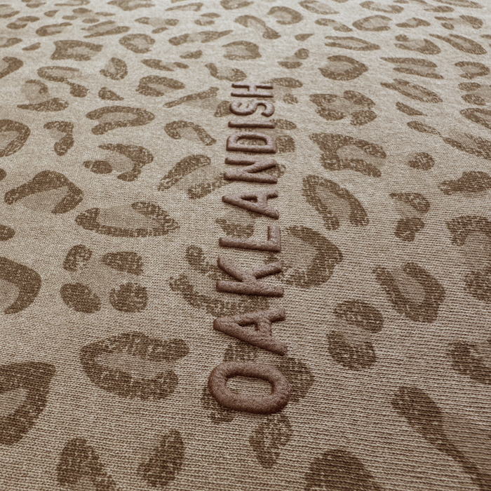 A close-up of heavyweight tan cotton with a brown leopard print and "OAKLANDISH" embossed vertically, perfectly matching the Oaklandish Classic Logo Tee’s color scheme.