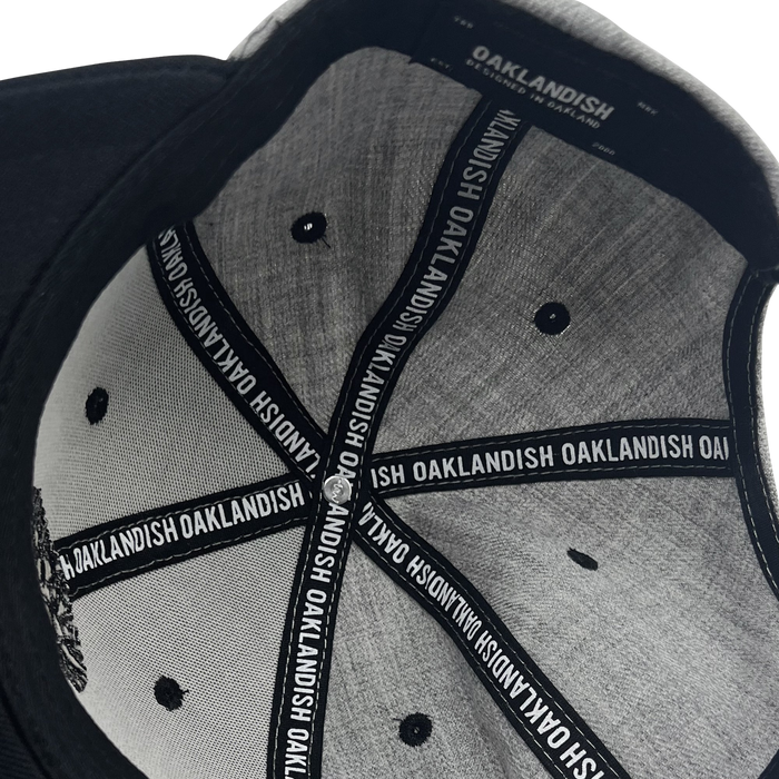 A close-up view of the Oaklandish Classic Snapback reveals its grey and black design. Labels attached to the inner seams feature the "Oaklandish" brand name in white text on a black background. The cap's interior stitching and structure, including fabric-covered ventilation holes, are clearly visible.