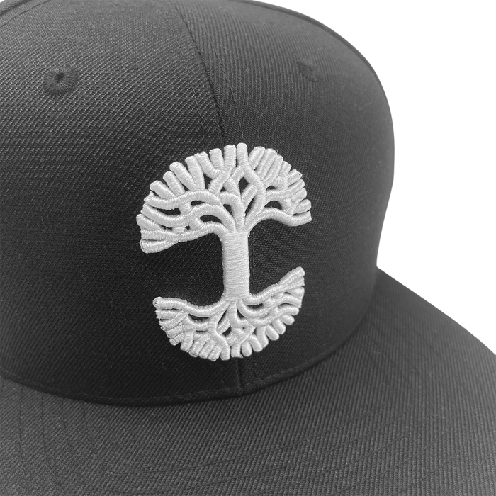 A close-up of the Oaklandish Classic Snapback cap by Oaklandish, highlighting a detailed white embroidered tree logo on its front panel. The design features a symmetrical tree with widespread branches at the top and mirrored roots at the bottom, creating an overall circular pattern. The background is plain white.