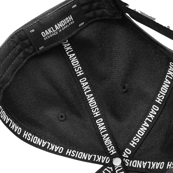A close-up shot of the inside of the Oaklandish Classic Snapback cap. The fabric on the inside is black, and the edges showcase intricate black and white stitching. The sweatband is adorned with the repeated text "OAKLANDISH." Additionally, a label states "OAKLANDISH DESIGNED IN OAKLAND EST. 2000." This stylish headwear exemplifies Oaklandish's finest craftsmanship.
