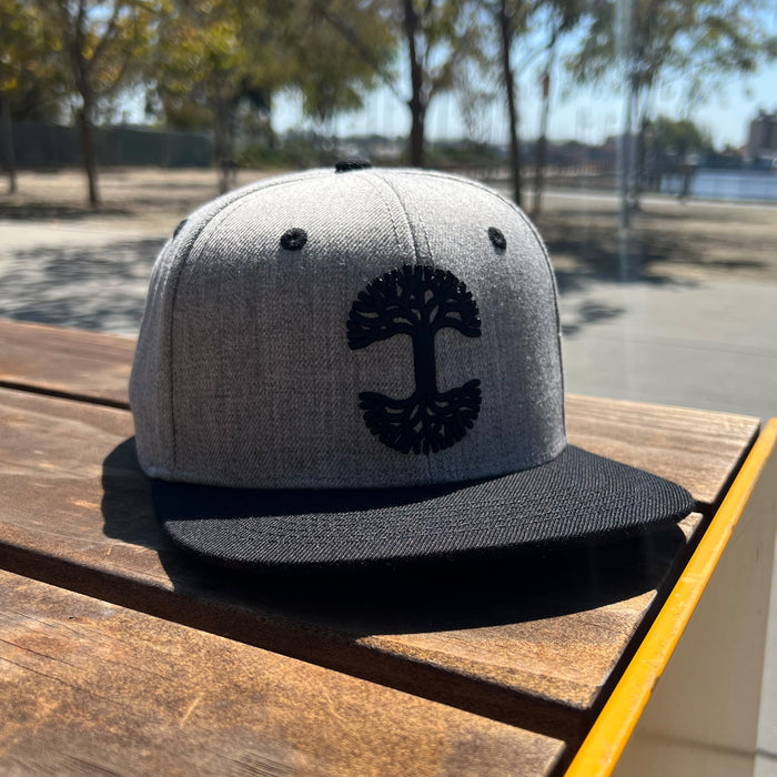 A gray and black Oaklandish Classic Snapback by Oaklandish, featuring a tree emblem on the front panel, is placed on a wooden table in an outdoor setting. Trees and a blurred view of a body of water are visible in the background, indicating a park or recreational area. The headwear is in clear focus under bright sunlight.
