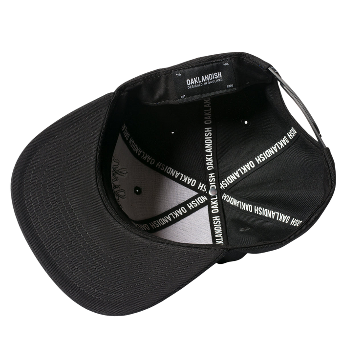 The Oaklandish Classic Snapback is shown from the underside, revealing its interior details. The inside features a black fabric lining adorned with white text that reads "OAKLANDISH" repeatedly along the seams. An adjustable snapback strap is visible at the back, and the inside tag also reads "OAKLANDISH.