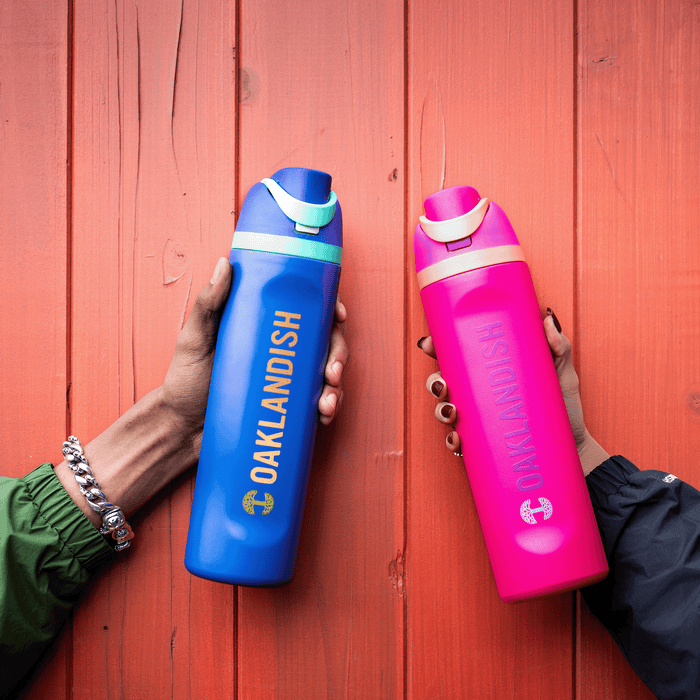 Two hands hold Oaklandish Colorful Bottles against an orange wooden background—one blue with "OAKLANDISH" in yellow, the other pink with "OAKLANDISH" in lighter pink. Both feature the Oaklandish tree logo and flip-top lids.