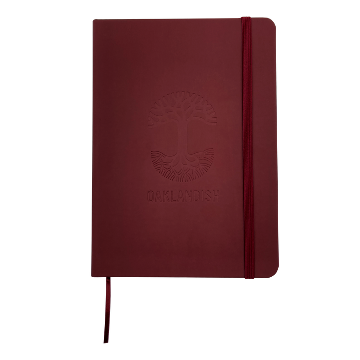 The Oaklandish Emblem Journal is a red 80-page lined notebook with a matching elastic band and ribbon bookmark. It features a debossed Oaklandish tree logo above the word "OAKLANDISH" on the PVC cover. The journal has rounded edges and a clean, minimalist design.