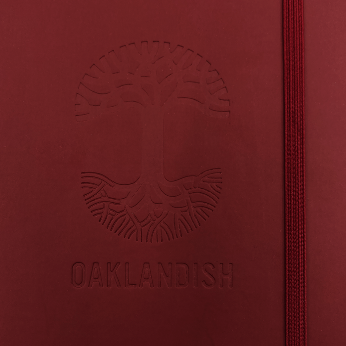 The Oaklandish Emblem Journal, produced by the brand Oaklandish, features a red PVC cover with a debossed tree logo and roots, along with the word "OAKLANDISH" beneath it. This 80-page lined notebook includes a red elastic band closure on the right edge. The design and text are subtly defined through the raised embossing.