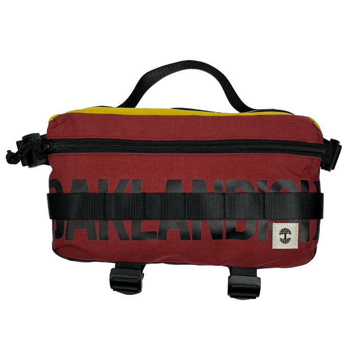 The Oaklandish Hip Bag is a rectangular, maroon waist bag featuring black zipper and buckle details for a premium look. The word "OAKLANDISH" is prominently displayed in black block letters across the front. A small white label with a black tree logo decorates the lower right corner, and a black handle on top adds to its unisex appeal.