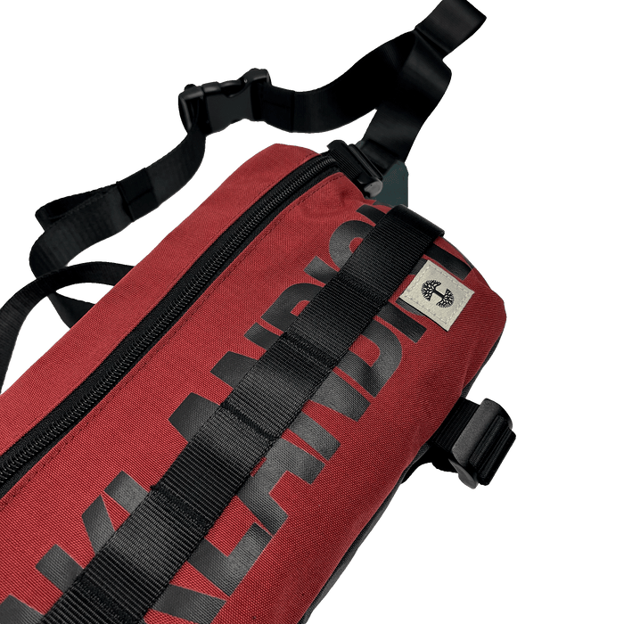 A close-up image of the Oaklandish Hip Bag, a red shoulder bag featuring black straps and a black zipper. The word "Oaklandish" is printed in bold black letters along the side of the bag. With its unisex appeal and premium details, including a small cream tag with the Oaklandish logo near the top, this versatile bag is positioned on a white background.