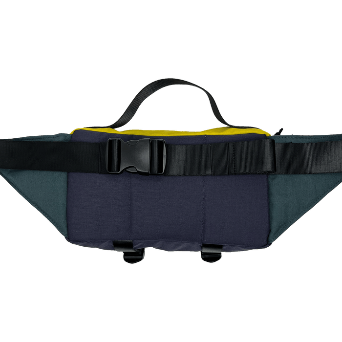 The Oaklandish Hip Bag by Oaklandish is a versatile waist pack featuring a combination of dark blue, grey, and yellow panels. It includes a top handle, an adjustable black strap with a plastic buckle closure, and two small strap loops at the bottom. Its compact design incorporates premium details, making it functional for carrying small items with unisex appeal.