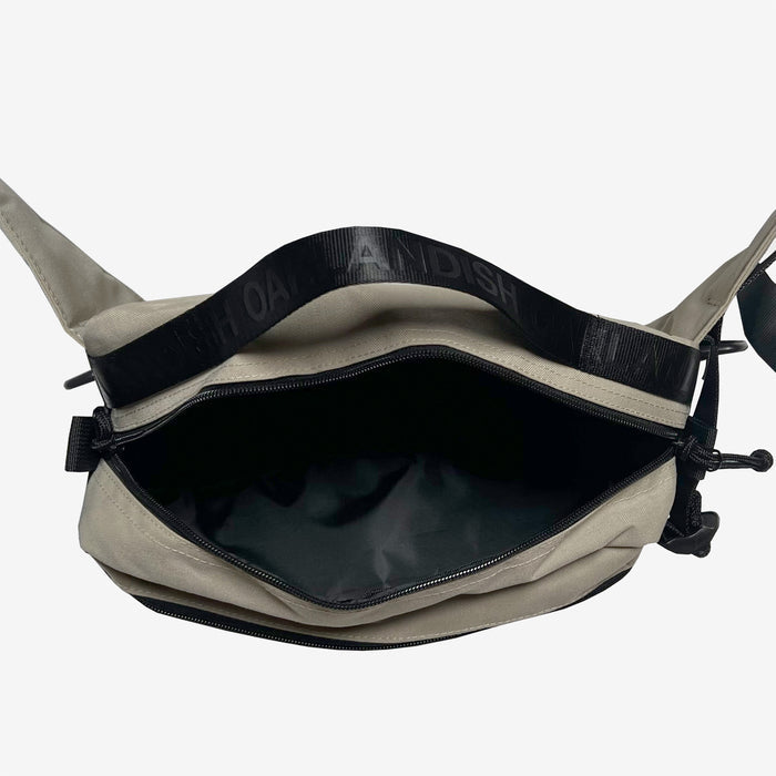 The Oaklandish Hip Bag is a beige crossbody featuring an open top view that reveals a spacious black inner compartment. The black strap has embossed text that partially reads "OAH AN DISH." This hip/shoulder bag from the Oaklandish brand showcases a zippered closure and minimalist design, set against a plain white background.