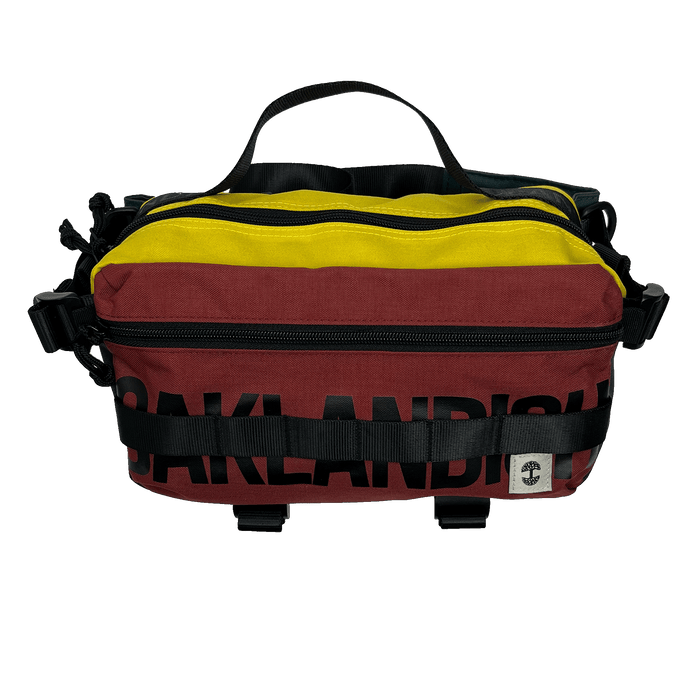The Oaklandish Hip Bag by Oaklandish is a small, colorful cross-body bag showcasing horizontal stripes in yellow, maroon, and black. The word "Oaklandish" is partially visible in large black letters on the maroon section. It features black zippers and adjustable straps for a versatile unisex appeal and is adorned with a premium white-and-black logo patch on the bottom right.
