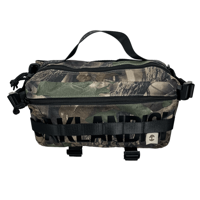 Front view of a camo patterned cross-body bag with front zipper, large black Oaklandish wordmark, and top handle.