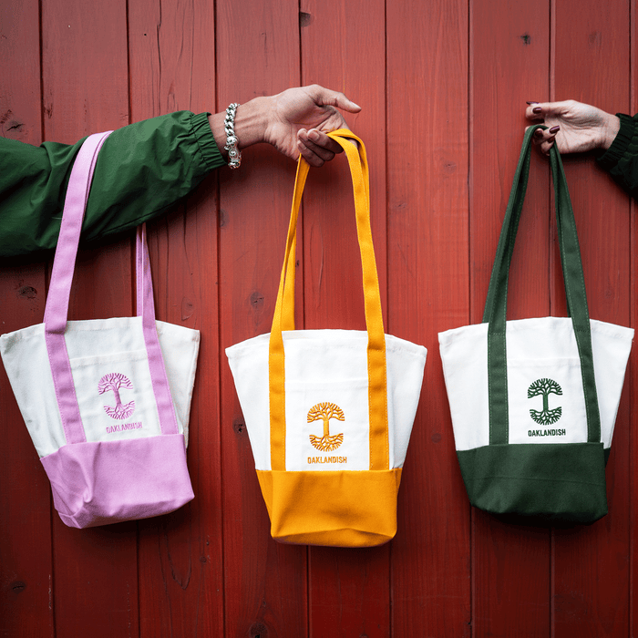 Two hands hold three Oaklandish Mini Grocery Totes against a red wooden background. The white bags have colored bases and straps—lavender, gold, and forest green—with a tree logo and “Oaklandish” on the front.